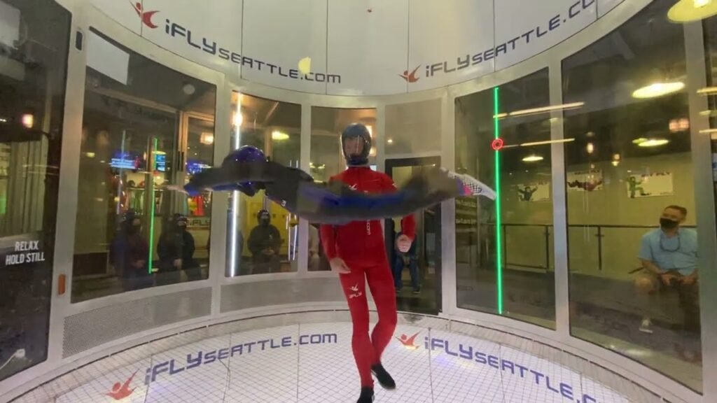 A person in a red flight suit and helmet experiences the sensation of free fall inside the glass wind tunnel at iFLY Seattle.