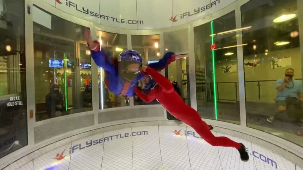 Two individuals enjoy vertical wind tunnel indoor skydiving at iFLY Seattle inside a circular glass chamber.