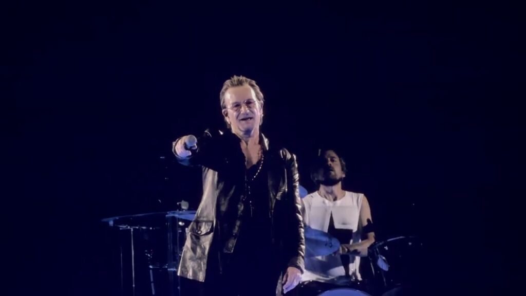 A performer on stage with a microphone, illuminated by blue stage lights, with a drummer in the background.