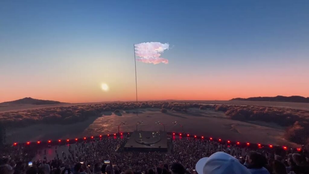 A large stage with red lighting sits in a desert landscape just before the sun sets below the horizon.