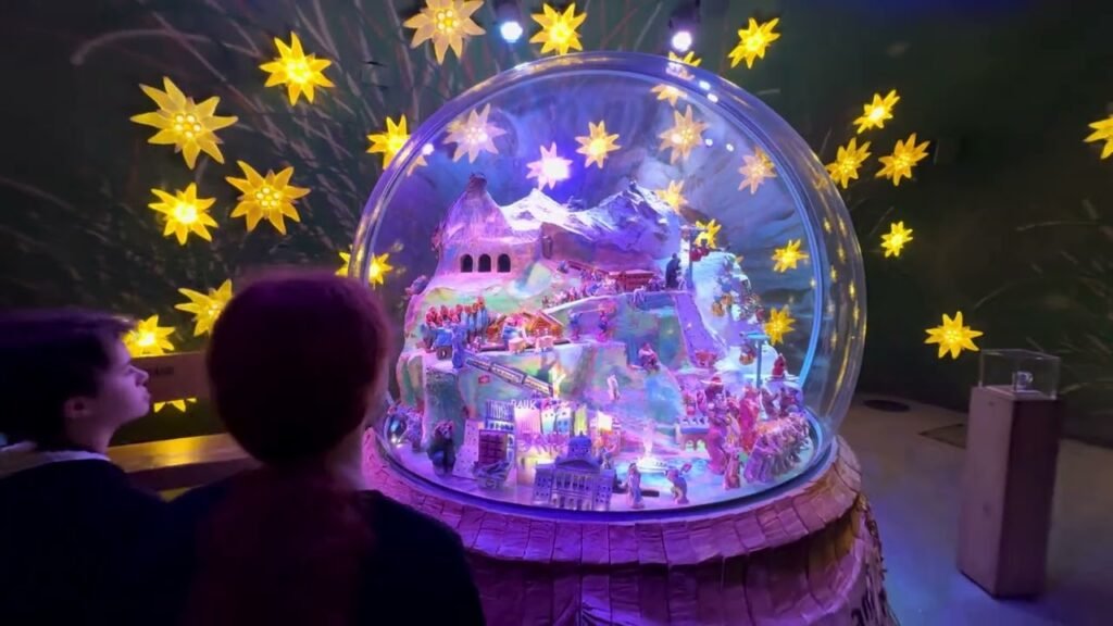 A large illuminated snow globe display featuring a glowing winter landscape surrounded by star-shaped lights and visitors observing the scene. Photo of a magical holiday display at night.