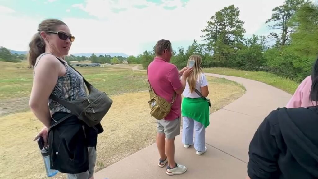 Kristie wearing a baby carrier stands beside a winding path surrounded by family and friends in an open field.