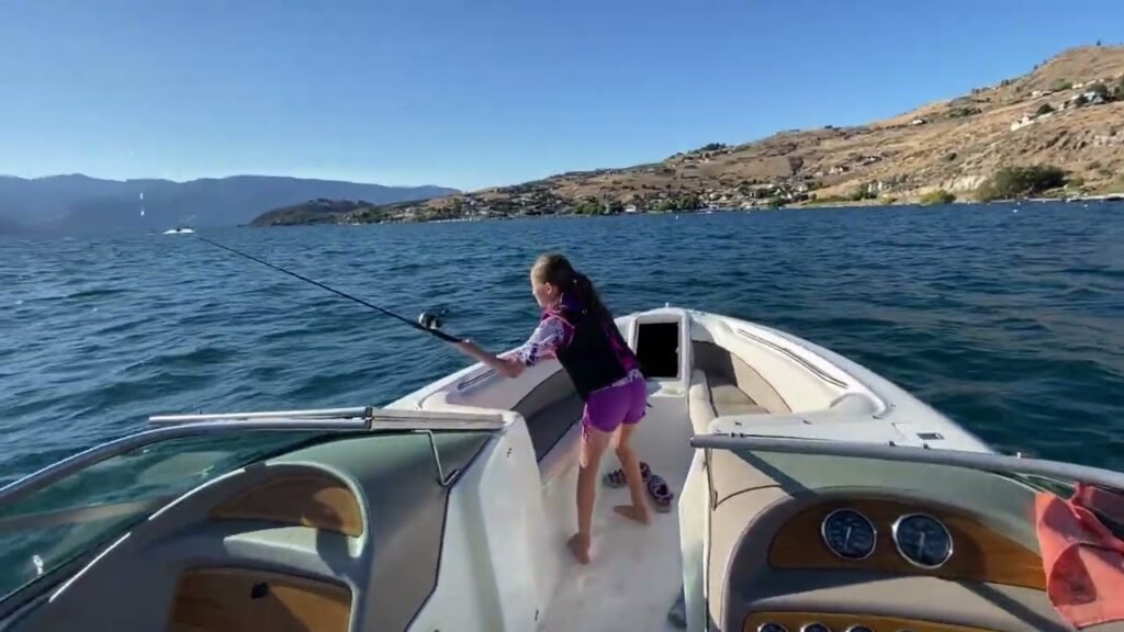 A child stands at the bow of a boat holding a fishing rod, casting a line into calm blue waters. The background features lush hills and a clear sky, typical of a day spent exploring the Pacific Northwest or a similar coastal region.