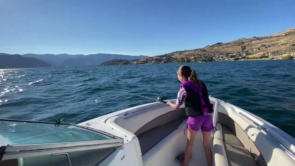 A young person stands on a boat, looking out over the blue ocean. The scene features clear water and scenic mountainous coastline.