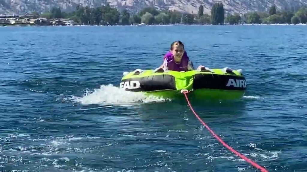 A person is riding a bright green ringo tow toy behind a boat on a large lake. [The water is blue with mountains and trees visible in the background.]