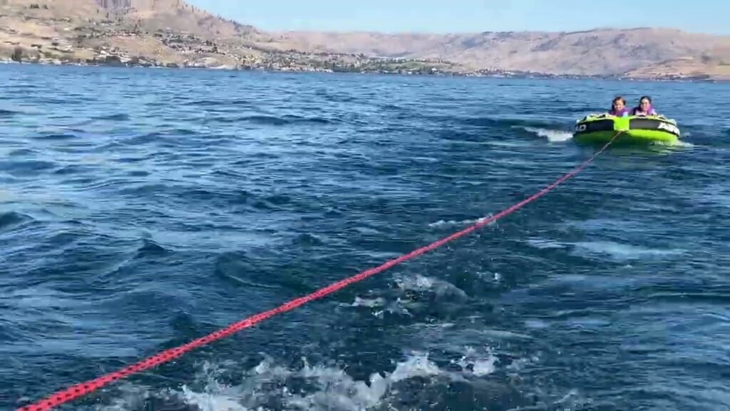 A green tube with two people in it being pulled across choppy blue water with a red tow rope.