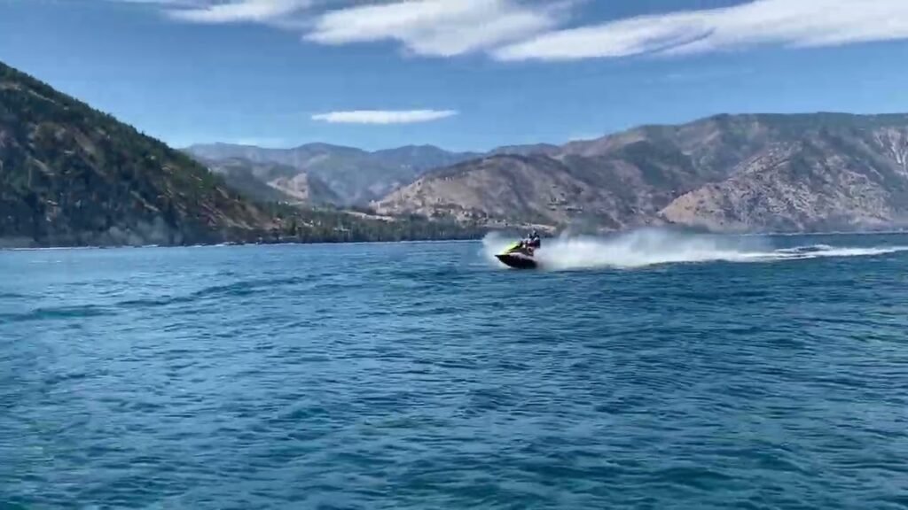 A jet ski speeds across a large lake surrounded by mountains. The water is deep blue with white spray behind the watercraft.