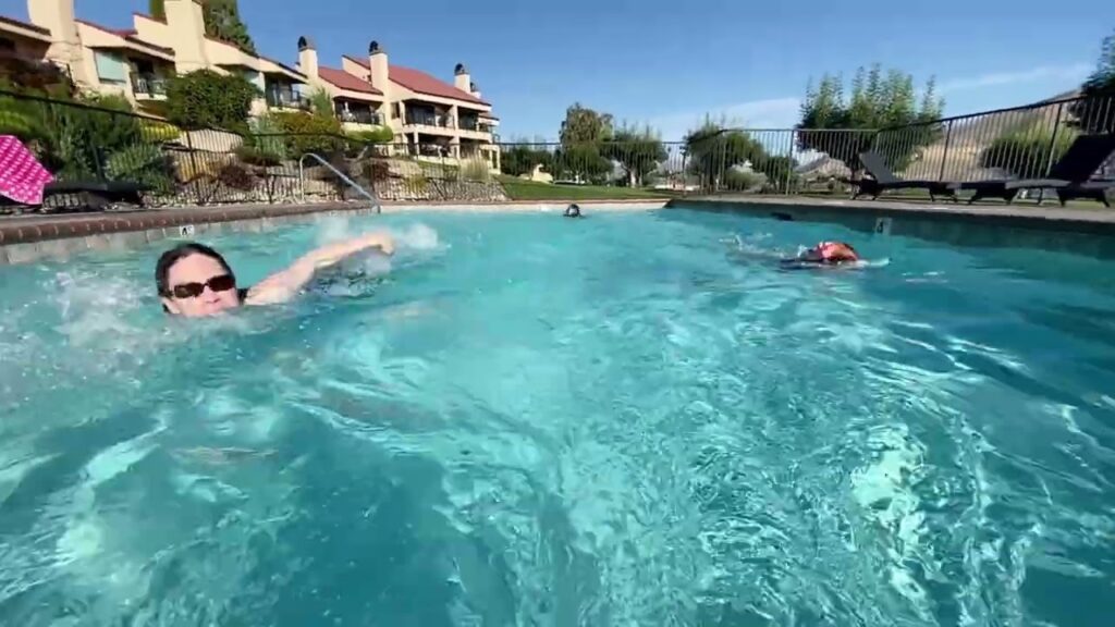 A person swims in a large resort pool with buildings in the background.