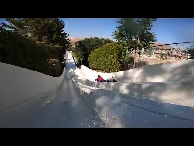 A person riding a waterslide with green trees and a blue sky in the background. The slide curves and bends through the garden settings.