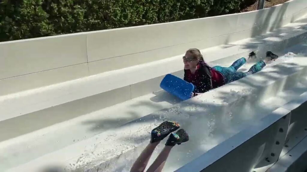 Person riding a white water slide with a blue float on a sunny day.