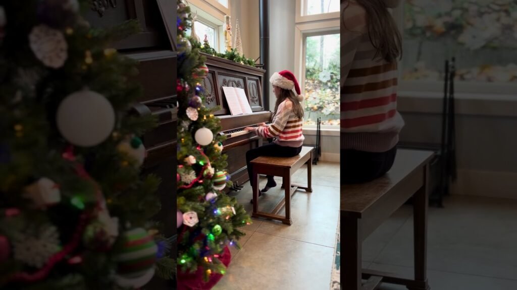 A child wearing a Santa hat plays the piano next to a decorated Christmas tree. Another individual is seated in the foreground listening.