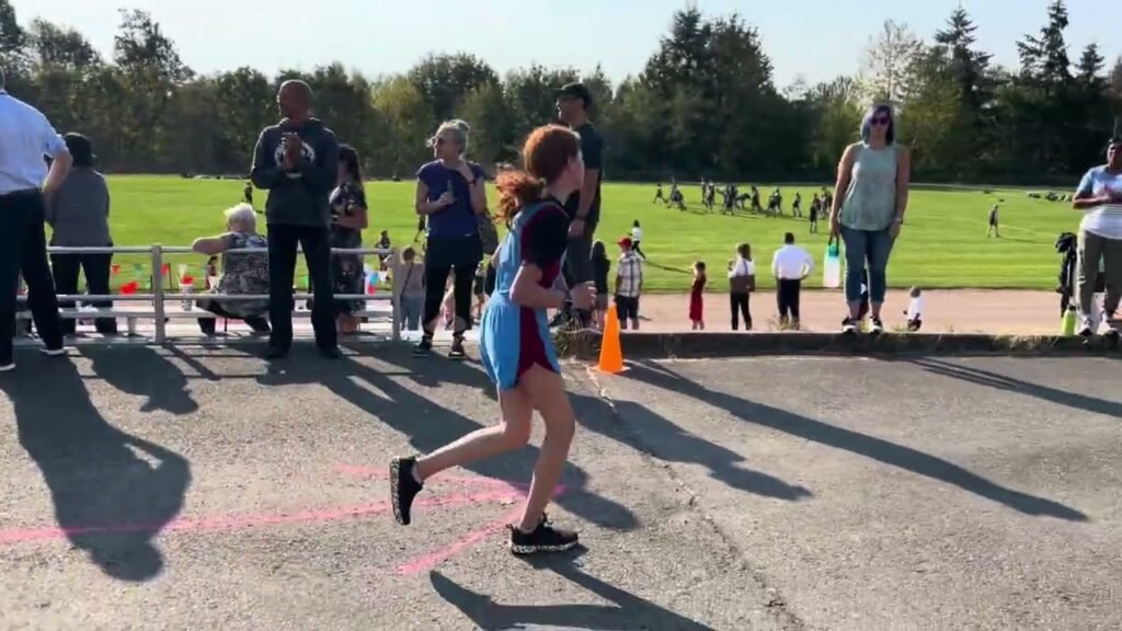 A young athlete in a blue and maroon uniform sprints toward the finish line at an outdoor track meet, with spectators standing behind a railing.