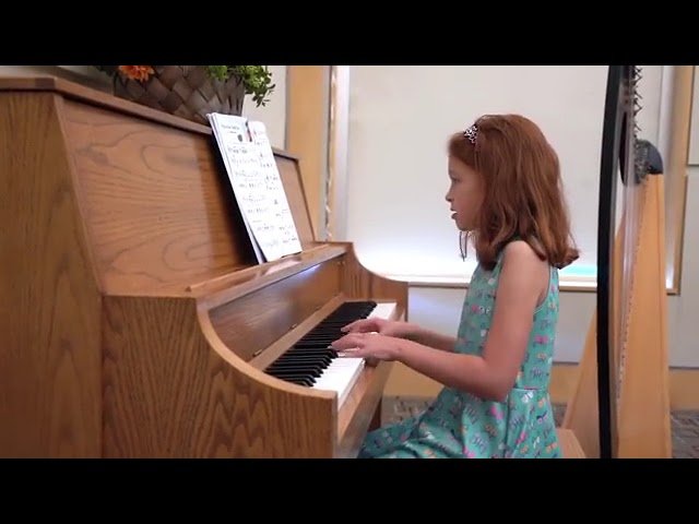 A red-haired pianist in a teal dress plays a wooden upright piano with focused attention. The image captures a moment of dedicated musical practice.
