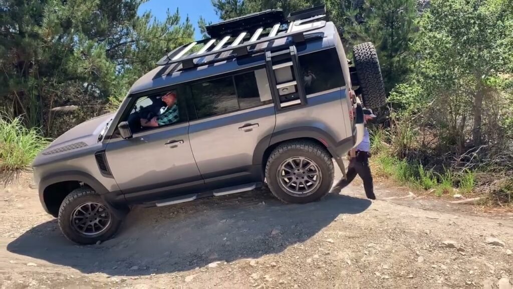 A modified Land Rover Defender navigates a steep, gravelly incline surrounded by dense greenery.