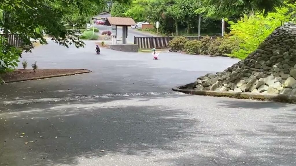 A wet, paved trail winding through a park with a stone pillar and a small shelter in the background, showcasing the beauty of the Pacific Northwest.