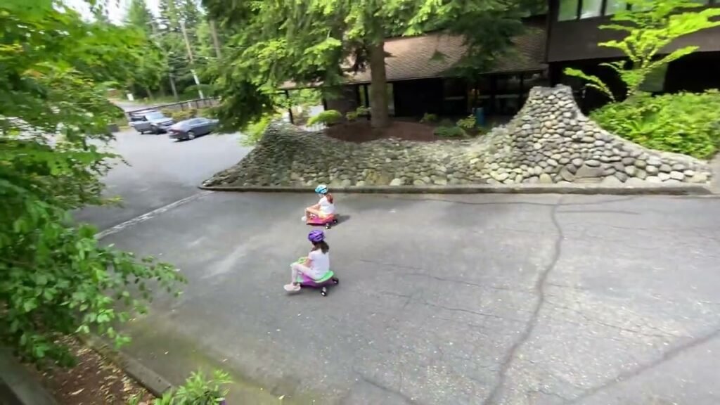 Two children stand on a driveway behind a house in a Pacific Northwest neighborhood, preparing to ride their skateboards.