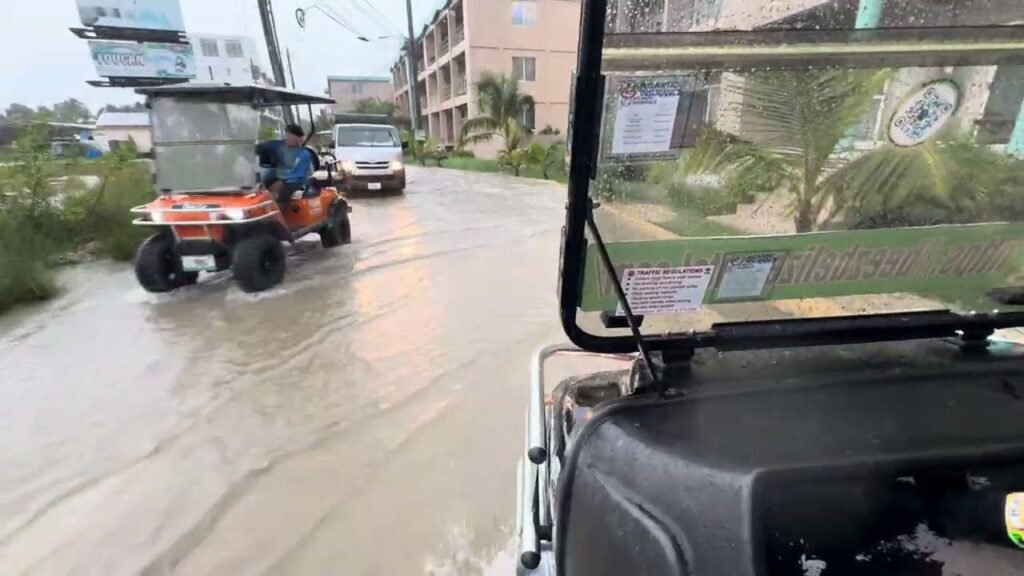 A utility vehicle drives through a flooded street alongside another vehicle in a tropical town.