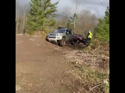 A rugged off-road truck navigating a dirt trail through the woods. A person in a high-visibility vest stands nearby.