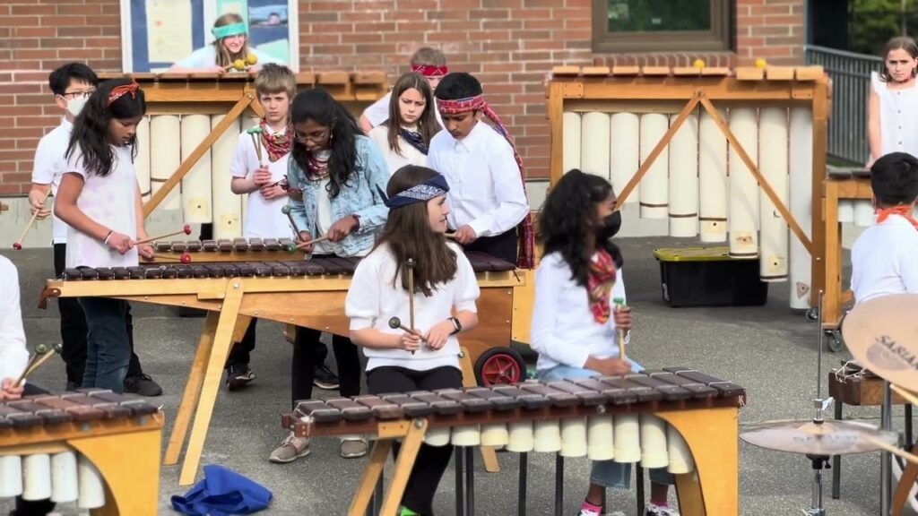 A group of young students wearing headbands and masks plays marimbas and xylophones in an outdoor courtyard, arranged near a large wooden musical instrument and a brick wall.
