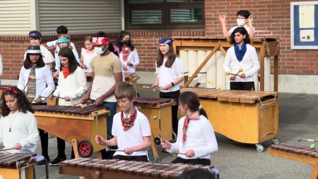 A group of children and adults play large wooden hand drums in an outdoor setting.