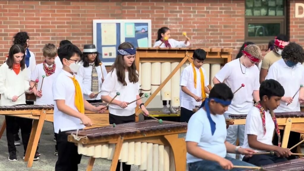 Students are playing hand drums while blindfolded in an outdoor group activity, wearing white shirts and colored scarves.