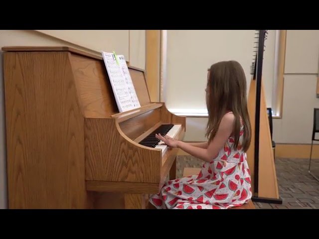 A person in a red and white dress sits at a wooden upright piano with open sheet music, with a stand visible in the background.