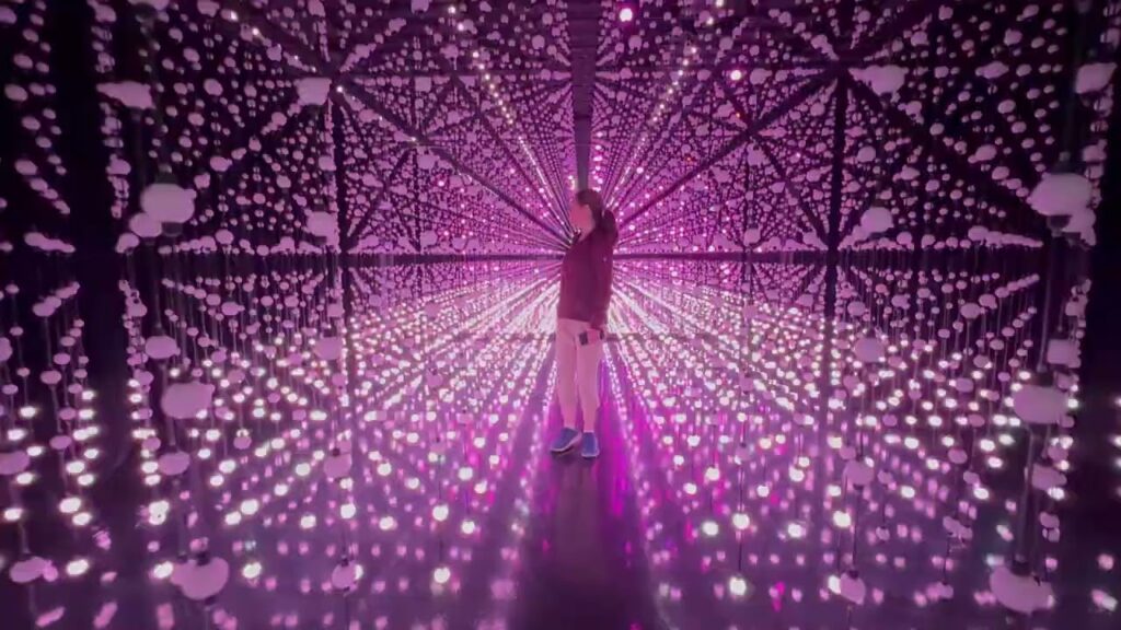A person stands in the center of an immersive purple light and mirror installation. The visual effect creates an infinite corridor with glowing dots and reflective surfaces.