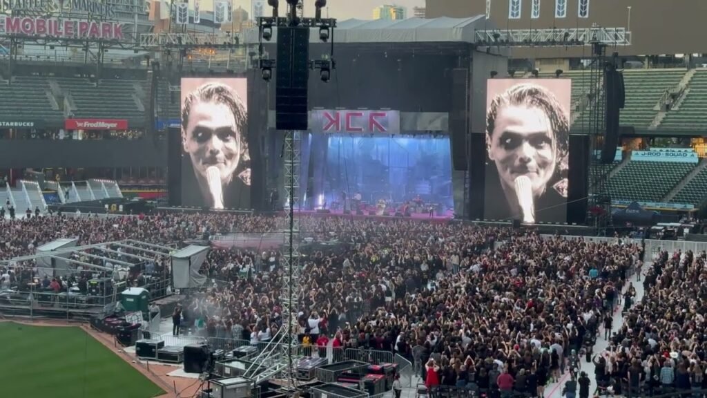 A large crowd fills a stadium in front of a concert stage, with a performer shown on giant video screens.