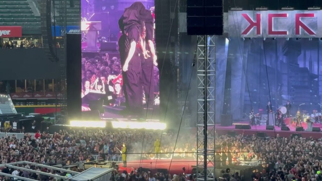 A large audience watches a stage performance under bright lights with a KCK banner visible above the stage.