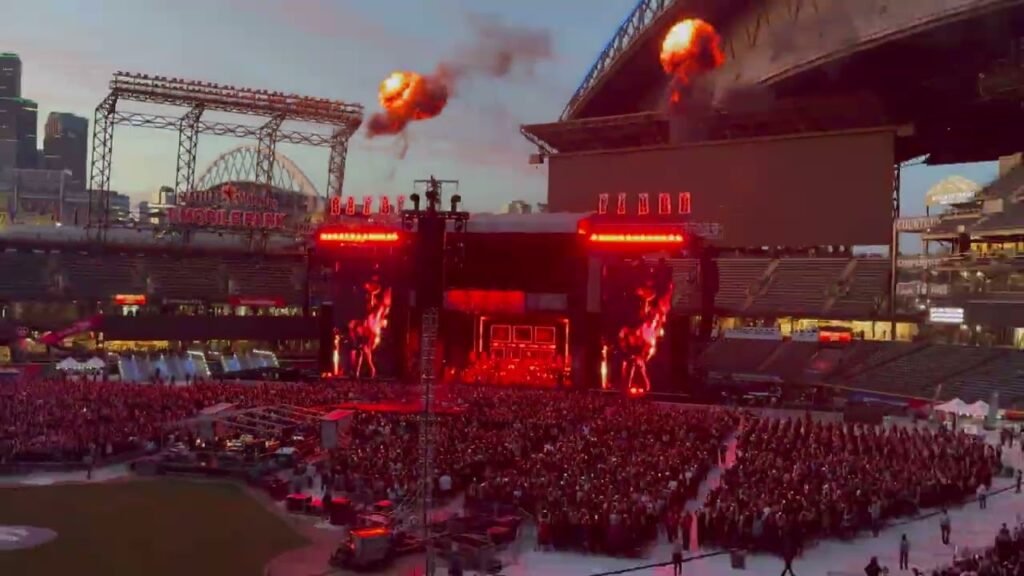 A large stadium audience gathers for an evening concert under red stage lighting and fireworks.
