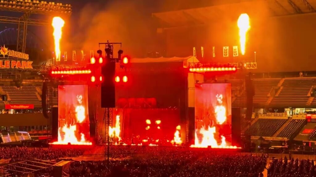 A concert stage illuminated by intense red lighting and dramatic fire pyrotechnics in a large stadium. Large screens display visuals before a massive crowd of attendees.