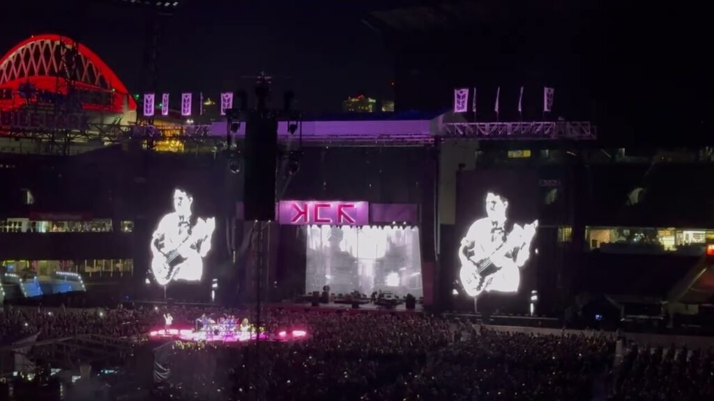 A stadium concert at night with performers on stage and large screens displaying a guitarist. Massive crowds fill the venue under vibrant lighting.