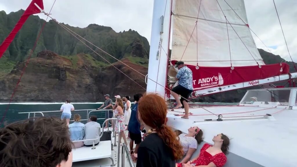 Passengers enjoy a sunny day on a red sailboat with a scenic coastal backdrop. A large sail with the name 'Andy's' is prominently displayed.