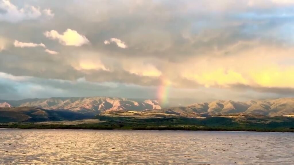 A scenic landscape showing a distant snow-capped mountain range under a cloudy sky at sunset. A faint rainbow arches near the mountains, with a large body of water in the foreground.