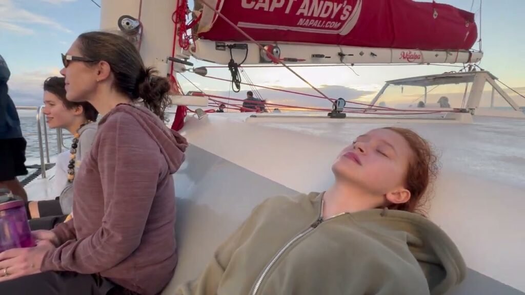 People relaxing on the deck of a sailboat, with one person lying down and others sitting near the water.