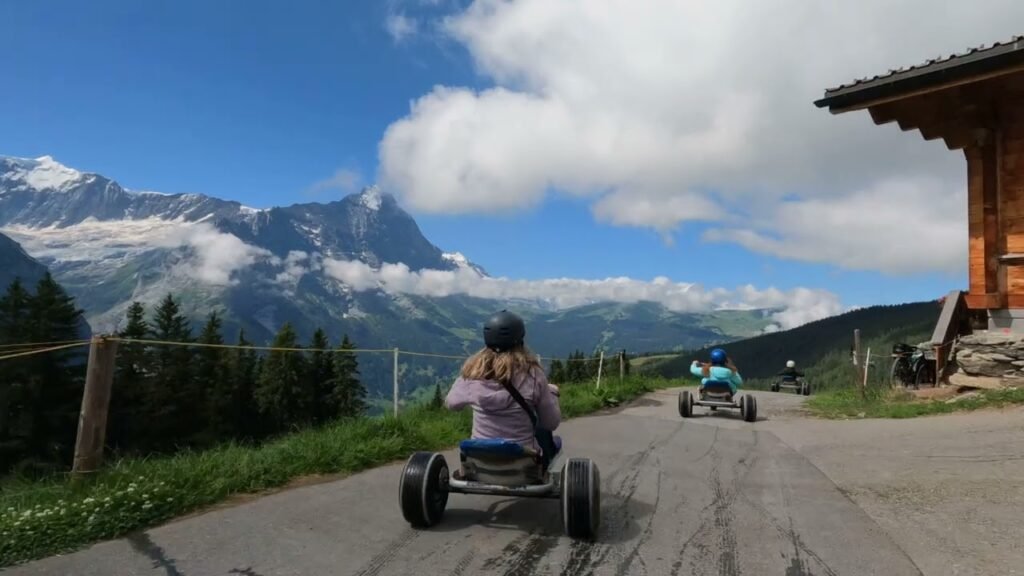 A person rides a gravity kart down a mountain road that passes a rustic wooden alpine hut, with snow-capped peaks visible in the background.