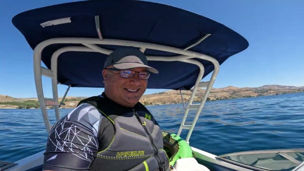 A person in boating gear stands on a boat with a blue canopy under clear skies.
