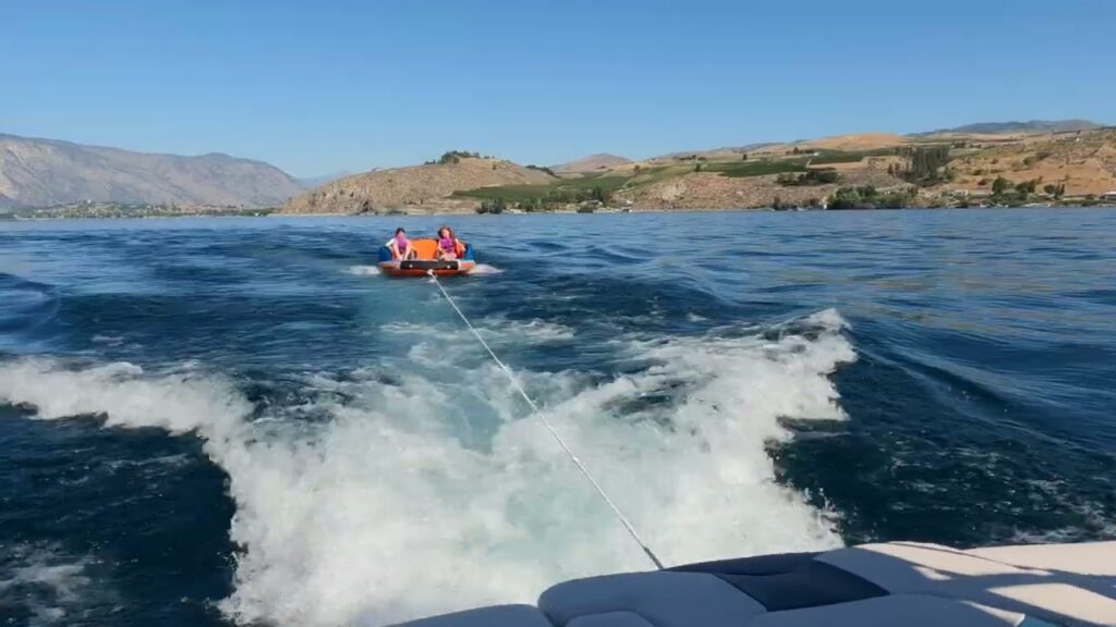 A speedboat towing an inflatable raft across a scenic body of water with hills in the background.