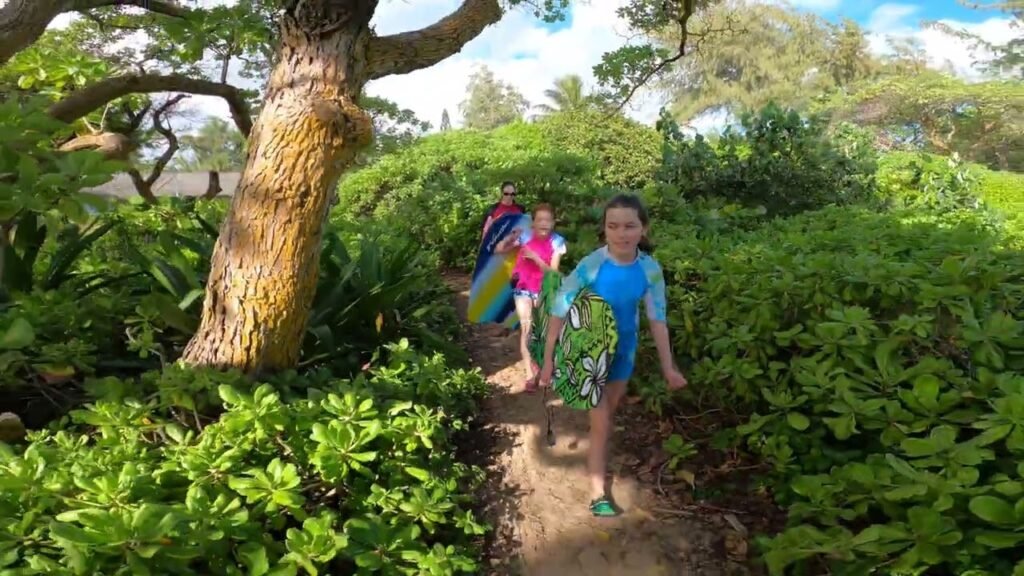 People walking on a dirt trail surrounded by dense green foliage and a large tree trunk in a garden setting.