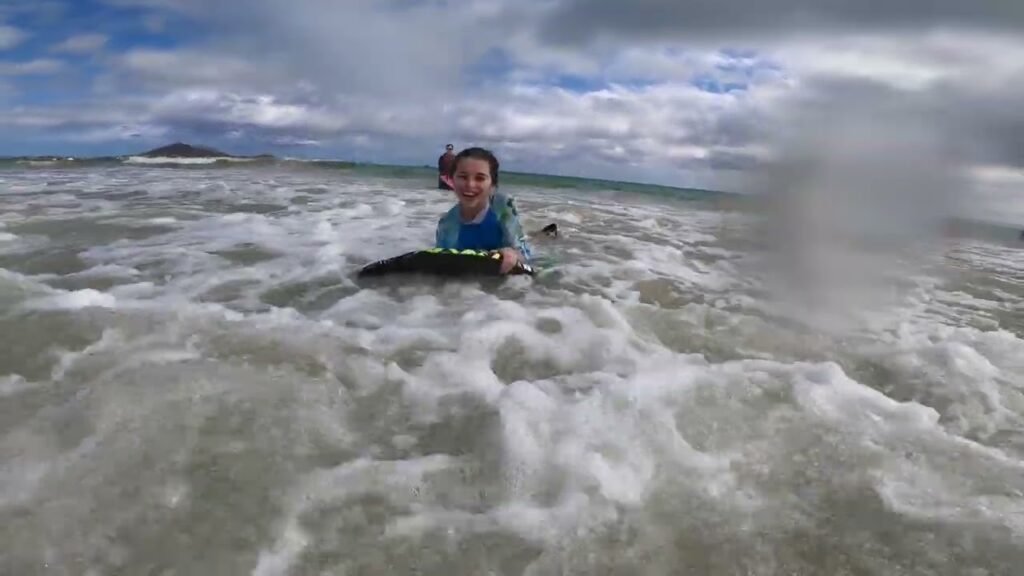 A person splashing in the ocean waves with a board. A small island is visible in the background under a cloudy sky.