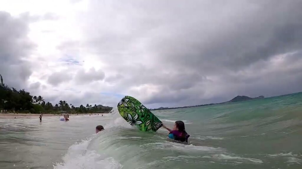 A person paddling a green surfboard in the ocean with a sandy beach in the background. The sky is overcast with soft, diffused lighting.