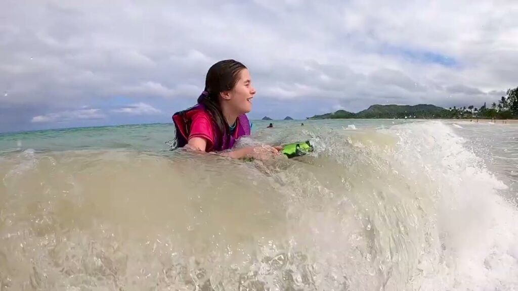 A person wearing a life jacket paddles a bodyboard while riding a wave in a tropical beach setting.