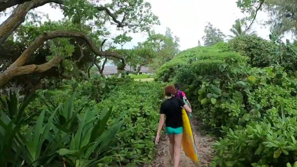 A traveler carrying a yellow surfboard walks along a narrow path through lush, dense tropical greenery.
