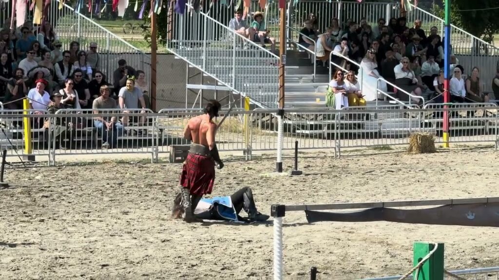 A performer in red pants stands over a fallen opponent in a sandy ring. Spectators watch from the bleachers in the background.