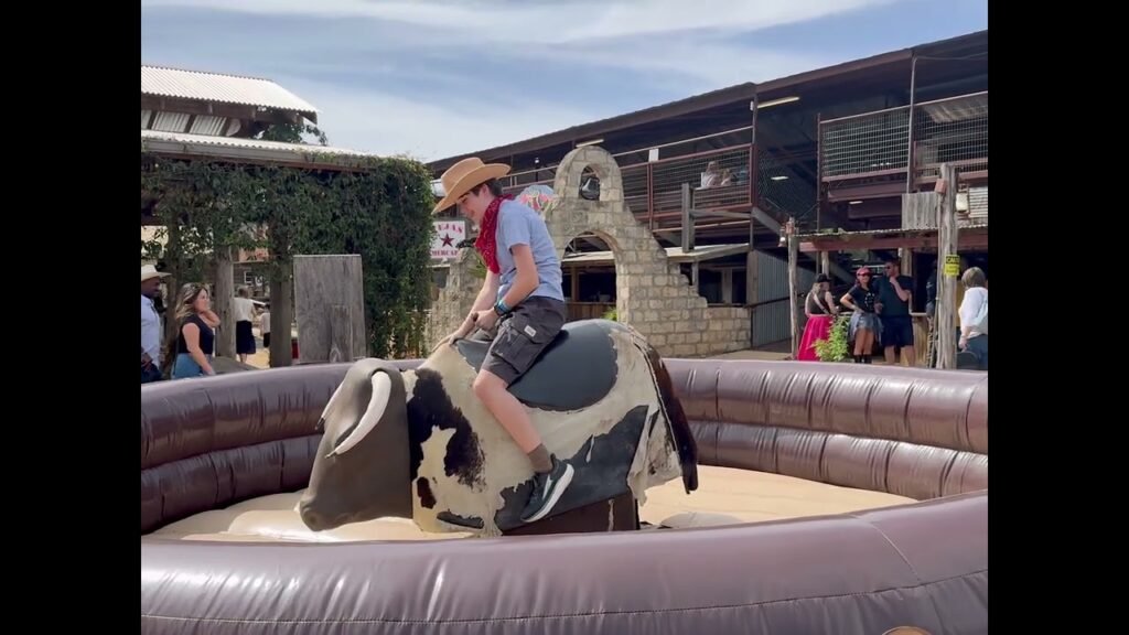 A person is riding a mechanical bull at an outdoor amusement park. The mechanical bull is situated in a padded arena surrounded by spectators.
