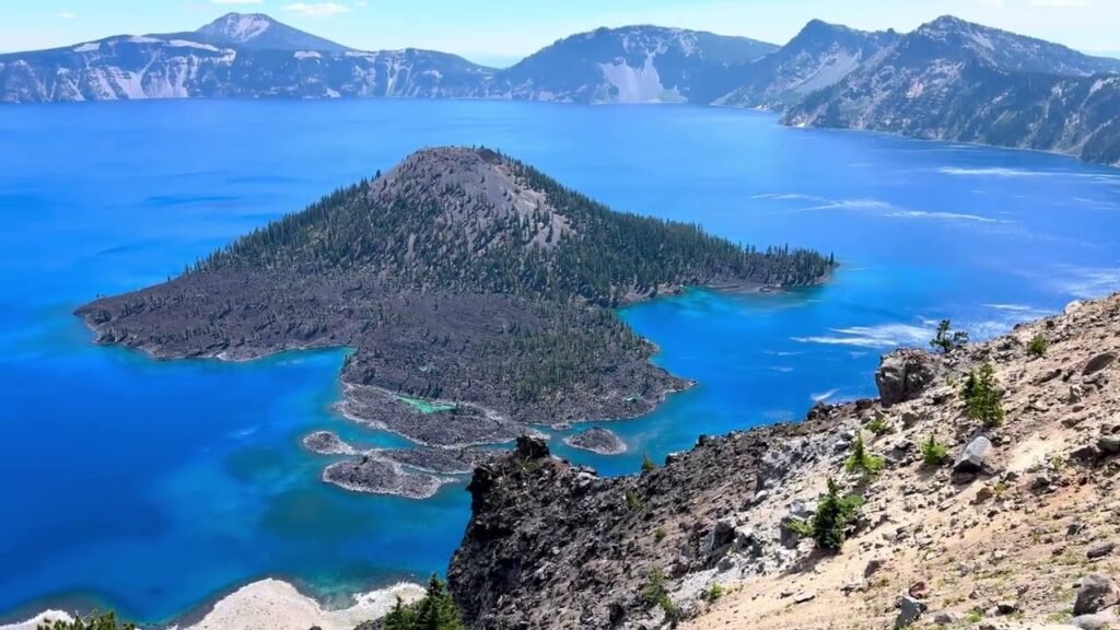 A scenic view of Crater Lake's deep blue waters and Wizard Island, surrounded by rugged cliffs and distant mountains.