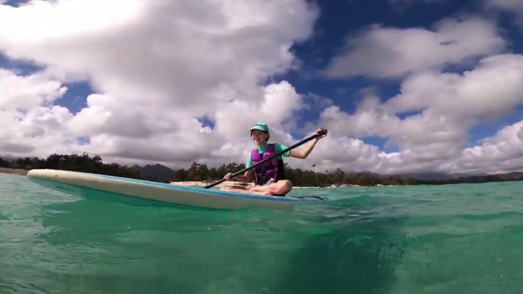 A person kayaking on a small boat in vibrant turquoise water with a cloudy sky and distant island in the background. A tranquil scene captured during a tropical adventure.