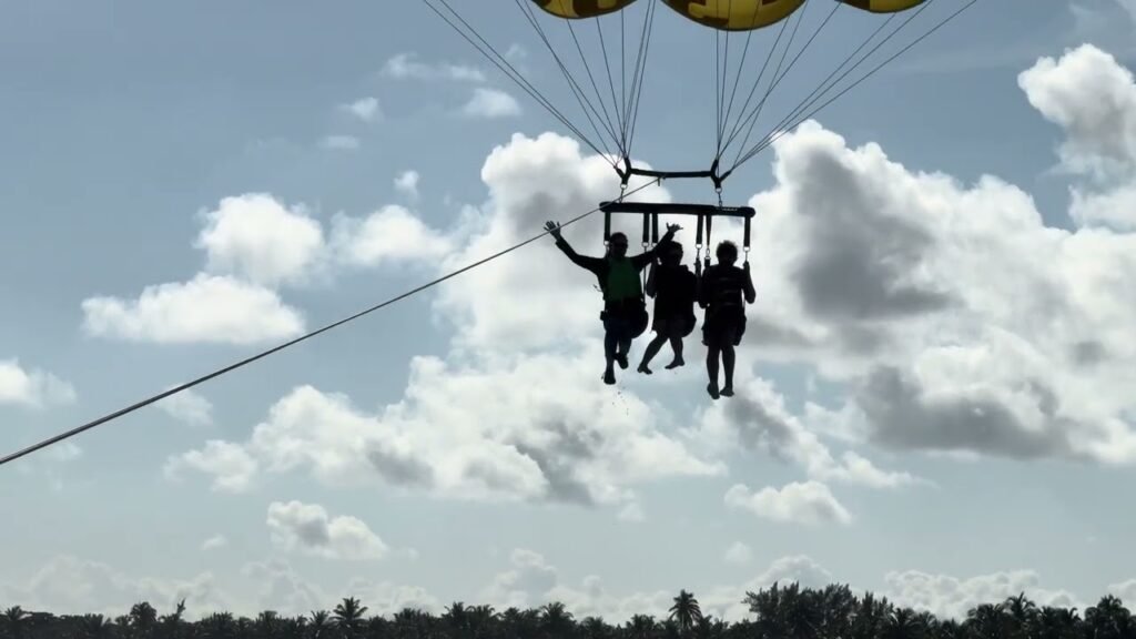 A group of people are seen hanging from a dangling platform attached to a parachute high above the ground under a bright, partly cloudy sky. A thin rope extends from the platform, with trees visible far below.