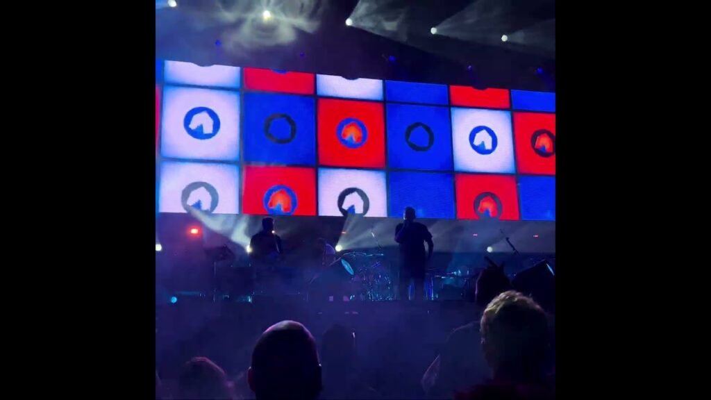 A large LED screen with a red and blue geometric pattern illuminates a concert stage above an audience.