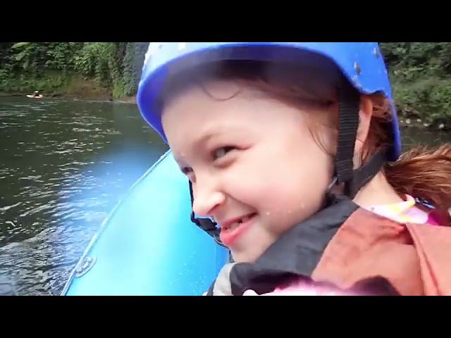 A smiling child wearing a blue helmet and life jacket enjoys a rafting trip on a calm river. The image captures close-ups of the person and the water, highlighting a fun outdoor family activity.